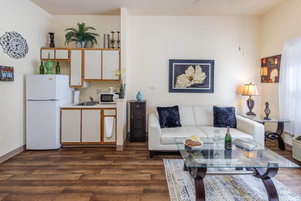 MiraLoma_INT_30_model Large - Vista Pointe at Mira Loma Inside of apartment showing kitchen area with refrigerator and microwave, living area with cozy couch, a coffee table with wine, book, and a snack. Artwork hangs on the walls. At the Vista Pointe at Mira Loma.