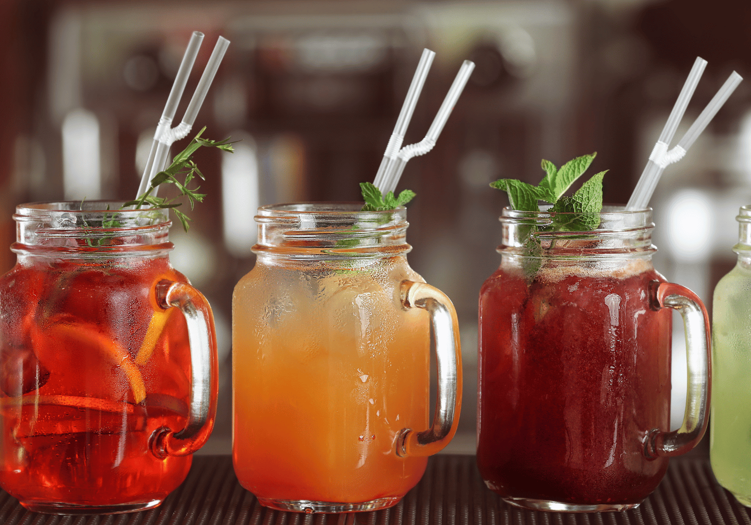 Three colorful iced drinks in mason jars with straws and mint garnish on a bar counter.