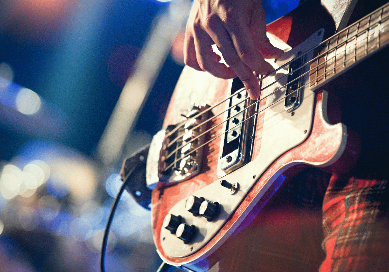 Close-up of a guitarist's hand picking strings on a worn red electric guitar on stage with blue bokeh lights in the background.